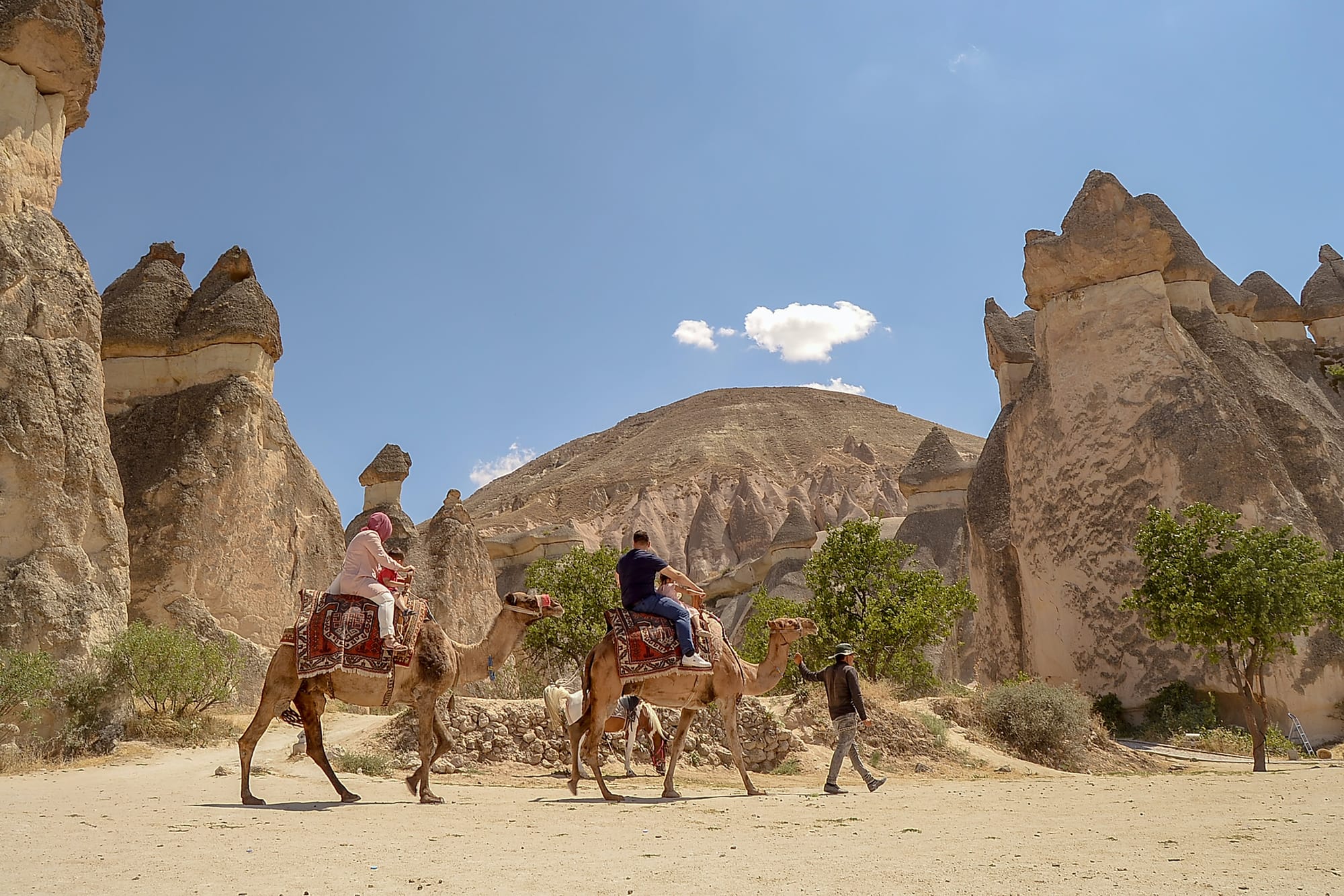 Tourists ride camels past the unique rock formations of Cappadocia under a clear sky.