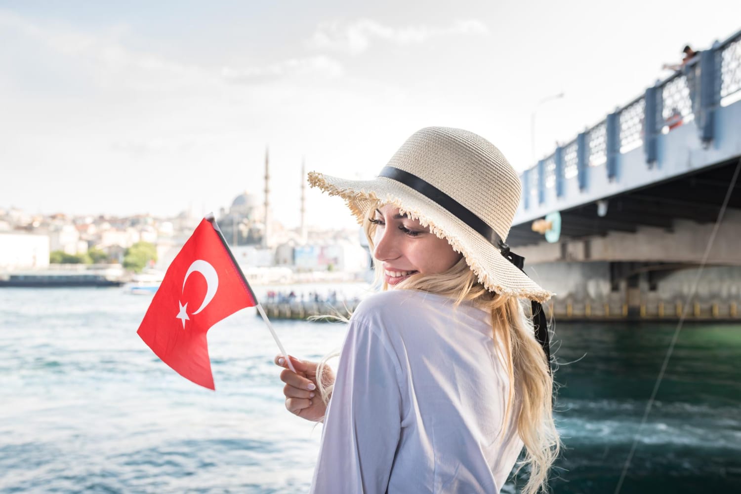 A woman in a sun hat smiles while holding a Turkish flag near a body of water, with a cityscape and a bridge visible in the background.
