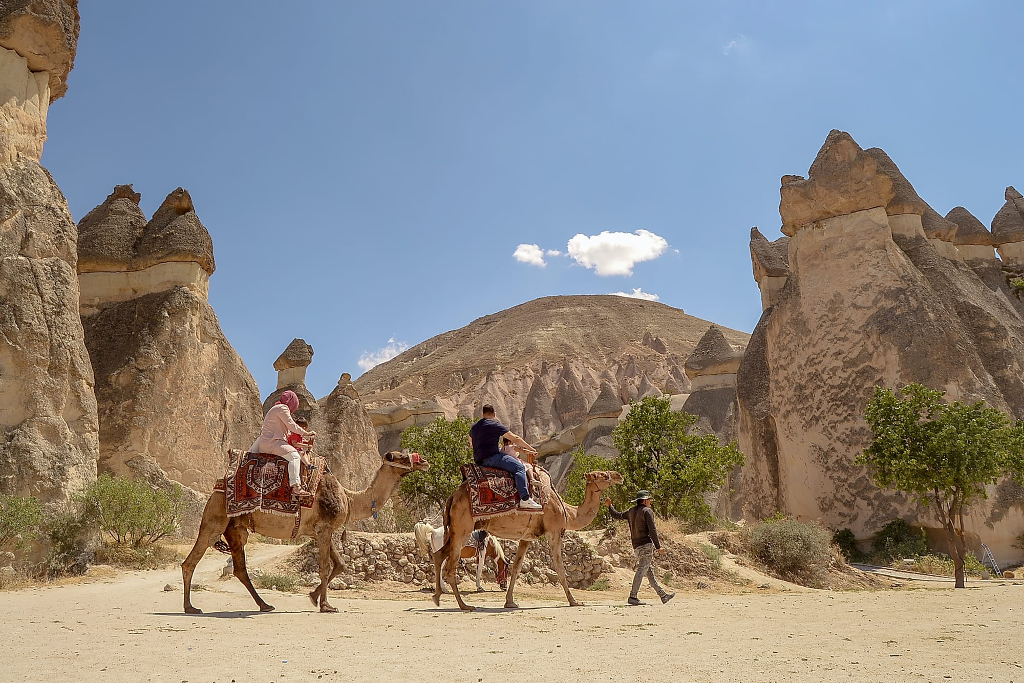 Tourists ride camels past the unique rock formations of Cappadocia under a clear sky.