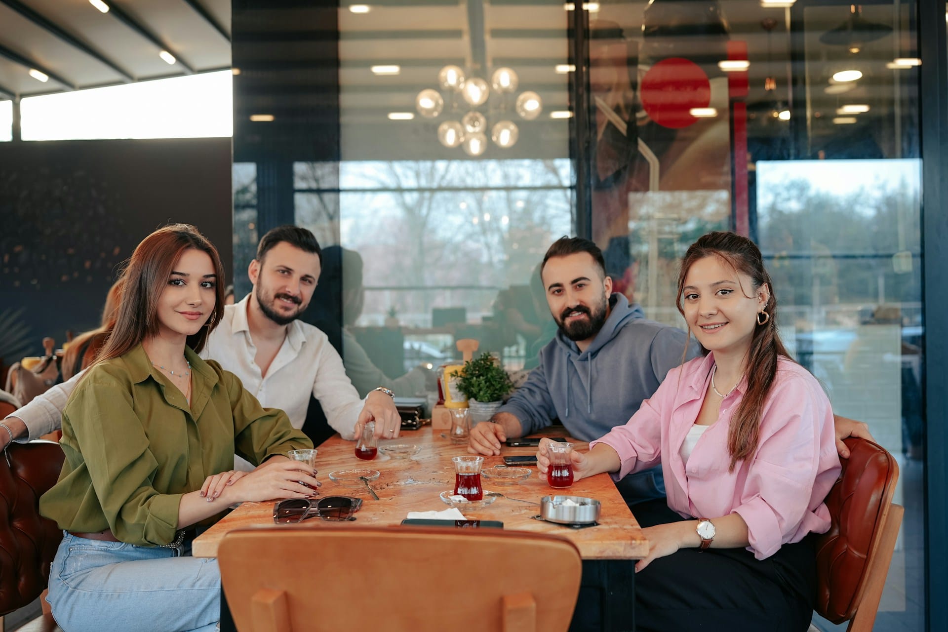 Four people sit around a café table, smiling at the camera and holding glasses of tea. The setting is bright and modern, with large windows and indoor plants in the background.